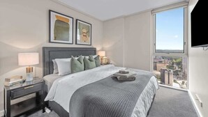 Ein modernes Hotelzimmer mit einem großen Bett, einem Nachttisch mit Lampe, Blick auf die Stadt durch ein großes Fenster und zwei gerahmten Kunstwerken an der Wand.
