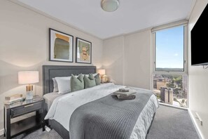 Ein modernes Hotelzimmer mit einem großen Bett, einem Nachttisch mit Lampe, Blick auf die Stadt durch ein großes Fenster und zwei gerahmten Kunstwerken an der Wand.