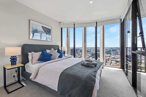 Ein modernes Schlafzimmer mit einem großen Bett, einem Nachttisch, Blick auf die Stadt und einem gerahmten Kunstwerk an der Wand.