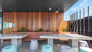 A modern outdoor seating area with wooden panel walls, concrete tables, and light-colored poufs. - Top Floor! 2bed/2bath/1car/pool ~ Views ~ CBD (Brisbane)