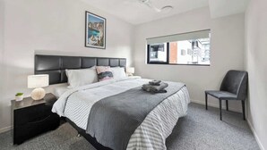 Quarto com cama, mesa de cabeceira, cadeira, ventilador de teto, janela e um quadro na parede.
