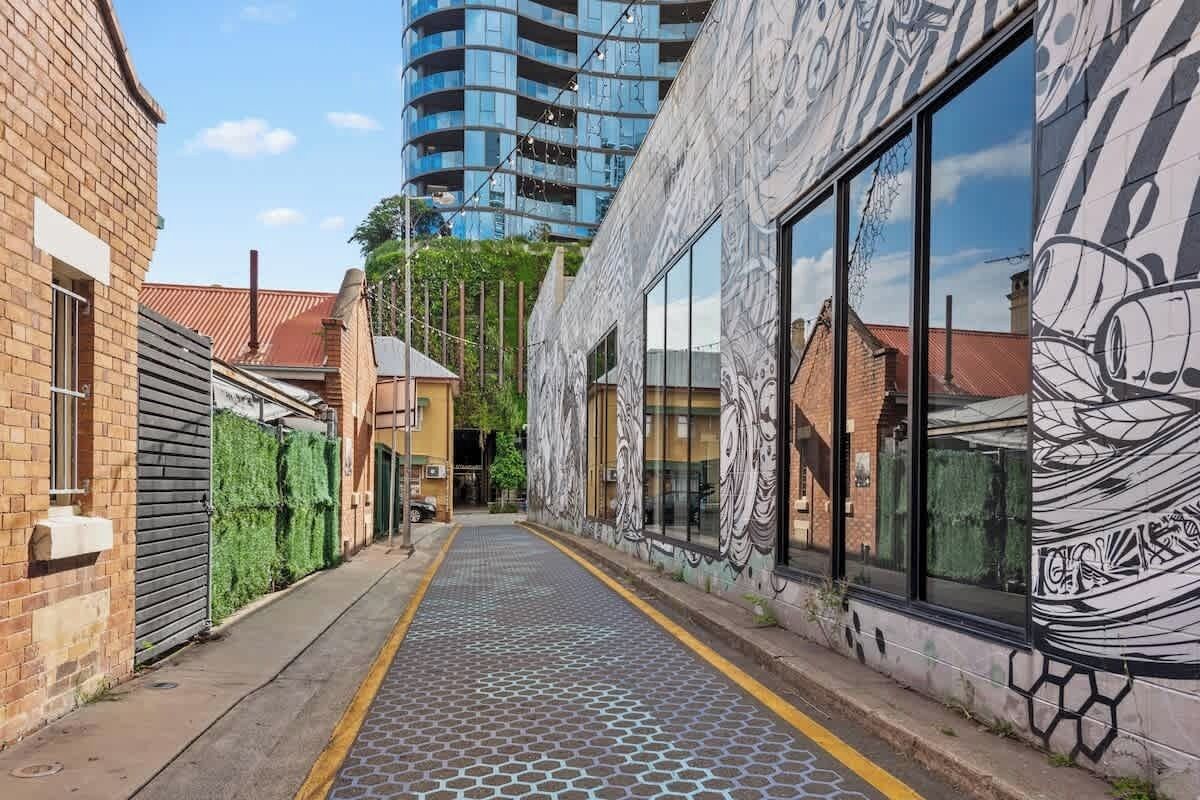 A narrow alley with a patterned pavement, a brick building with a green hedge, and a modern building with large windows and a mural.