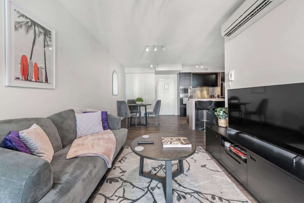 A modern living room with a sofa, coffee table, and a flat-screen TV. There is a dining area with a table and chairs, and a kitchen with a microwave and stove.