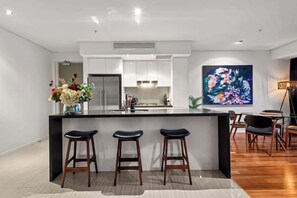 A modern kitchen with a central island, bar stools, and a large abstract painting on the wall. - Skyhigh Executive! ~ 2bed/2bath/1car/views ~ CBD (Brisbane)