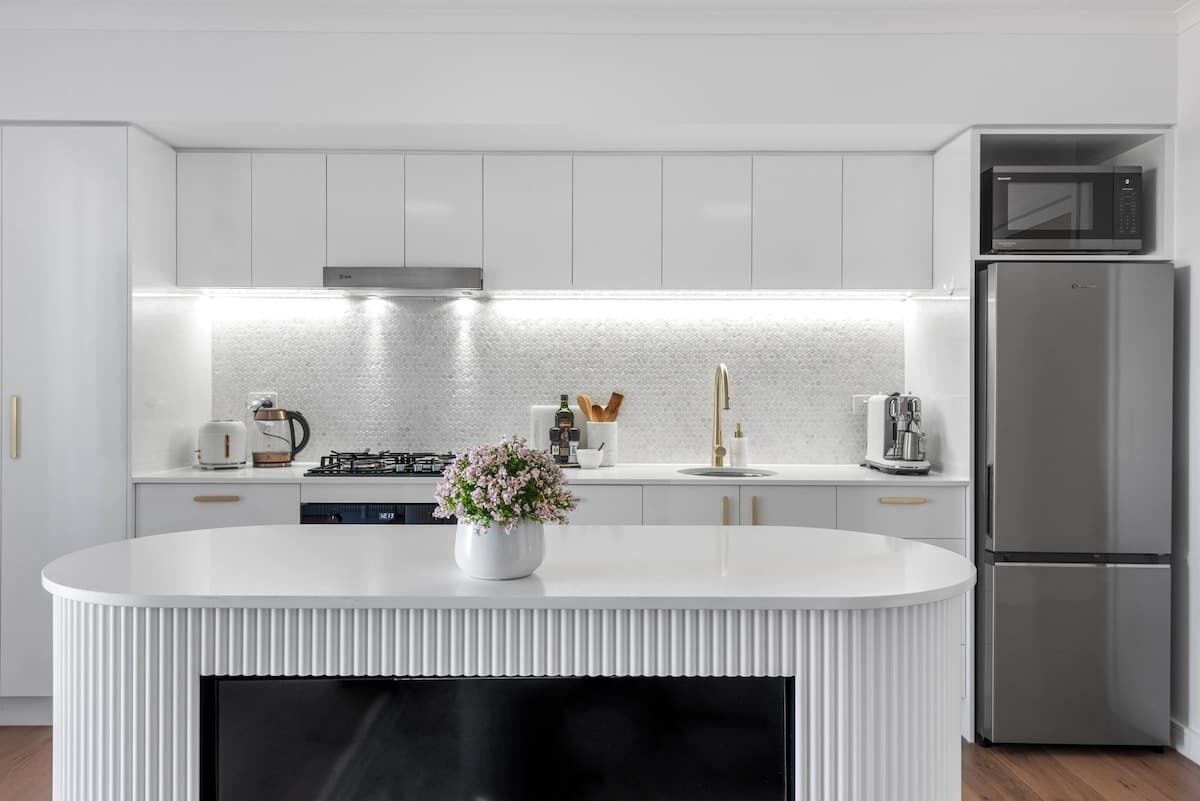 Une cuisine moderne avec des meubles blancs, un réfrigérateur en acier inoxydable et un îlot central doté d’un écran noir.