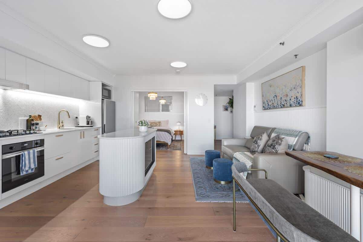 A modern kitchen with white cabinetry, a central island, and a dining area with a sofa and blue ottomans.