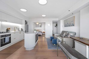 A modern kitchen with white cabinetry, a central island, and a dining area with a sofa and blue ottomans. - Sweet! 1bed, 1bath, 1car, Views ~ CBD (Brisbane)
