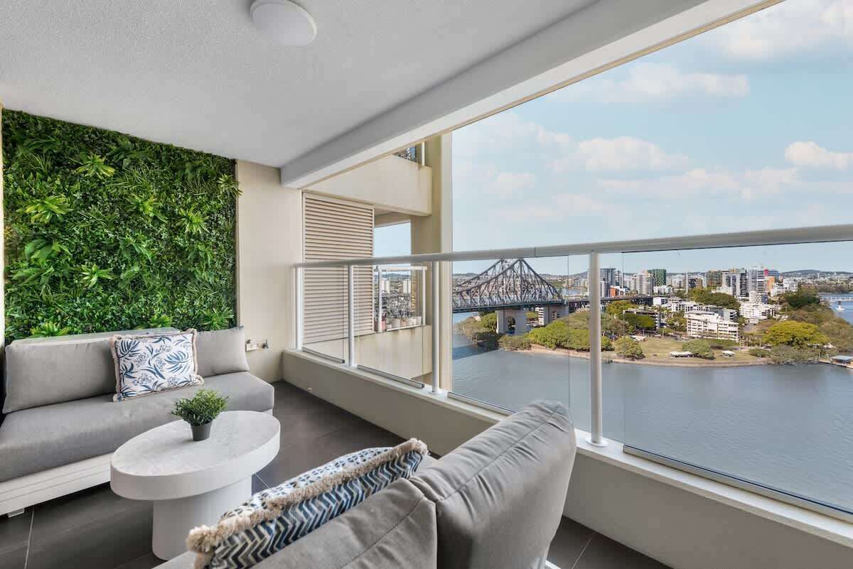Un balcon moderne offrant une vue sur un pont et sur l’eau, avec un canapé gris et une table ronde agrémentée d’une plante.