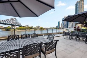 An outdoor seating area with tables and chairs, overlooking a river and a bridge, with city buildings in the background. - Sweet! 1bed, 1bath, 1car, Views ~ CBD (Brisbane)