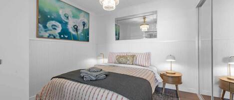 A bedroom with a bed, a chandelier, a painting, a mirror, and a bedside table.