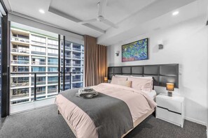 Ein modernes Hotelzimmer mit einem großen Bett, einem Nachttisch, einem Deckenventilator, einem Gemälde und Blick auf ein Hochhaus.