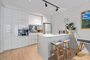 A modern kitchen with white cabinetry, a central island with stools, a clock on the wall, and a painting above the island. - South Bank Living! ~ 1bed, 1bath, 1car, Balcony (Brisbane)