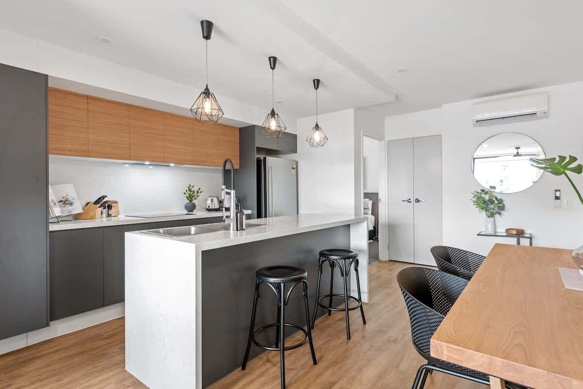 Une cuisine moderne dotée d’un îlot central, de tabourets de bar, de luminaires suspendus et d’un coin repas avec une table en bois.
