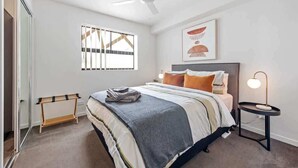 Un dormitorio con cama, ventilador de techo, mesita de noche con lámpara, ventana con persianas y un perchero para toallas.