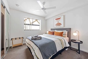 Ein Schlafzimmer mit einem Bett, einem Deckenventilator, einem Nachttisch mit Lampe, einem Fenster mit Jalousien und einem Handtuchhalter.