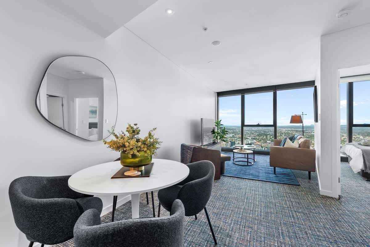 A modern hotel room with a dining area, a sofa, a TV, and a large window offering a city view.