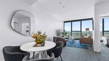 A modern hotel room with a dining area, a sofa, a TV, and a large window offering a city view.