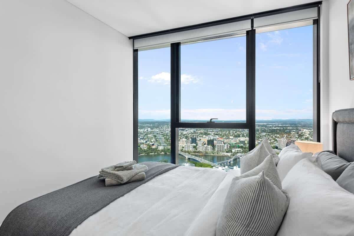 A bedroom with a large window, a bed with pillows, and a view of a cityscape.