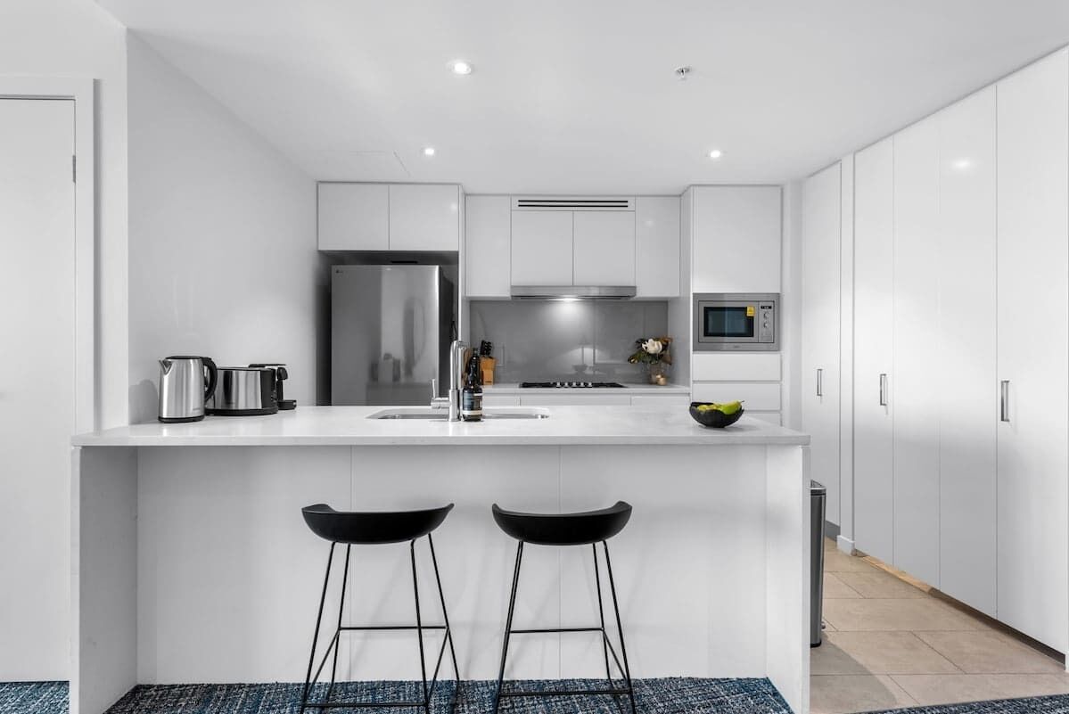 A modern kitchen with white cabinetry, a stainless steel refrigerator, a microwave, and a sink. There are two black bar stools at the island.
