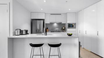 A modern kitchen with white cabinetry, a stainless steel refrigerator, a microwave, and a sink. There are two black bar stools at the island.