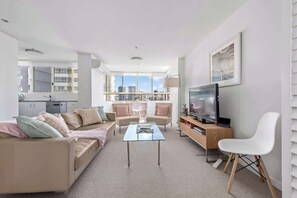 Un salon moderne avec un canapé, une table basse, un téléviseur et une vue sur la ville grâce à de grandes fenêtres.