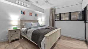 Un dormitorio con cama, mesitas de noche, aire acondicionado, ventilador de techo y ventana con persianas.