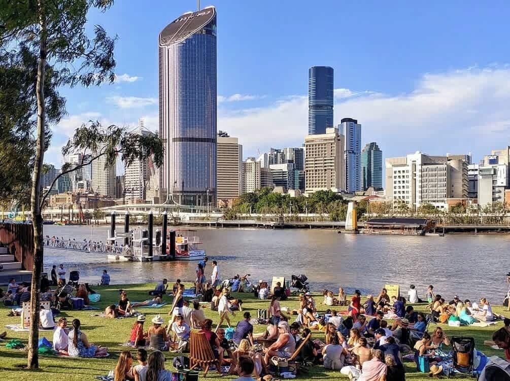 A riverside park with people relaxing on grass, a bridge, and a city skyline with modern buildings in the background.