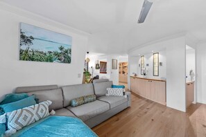 A living room with a grey sofa, a painting of a beach, and a kitchen area with wooden cabinets. - Point Peninsula - 2bed-2bath-car-pool - Teneriffe (Brisbane)