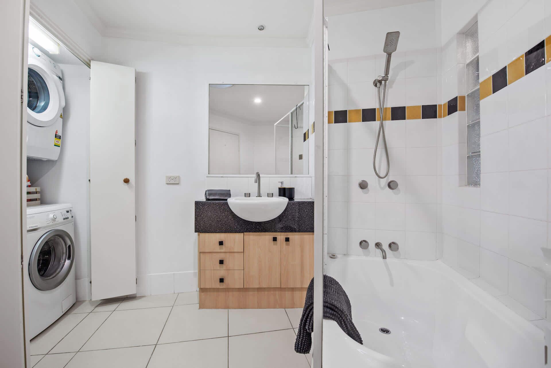 A modern bathroom with a washing machine, a sink with a granite countertop, a mirror, and a shower area.