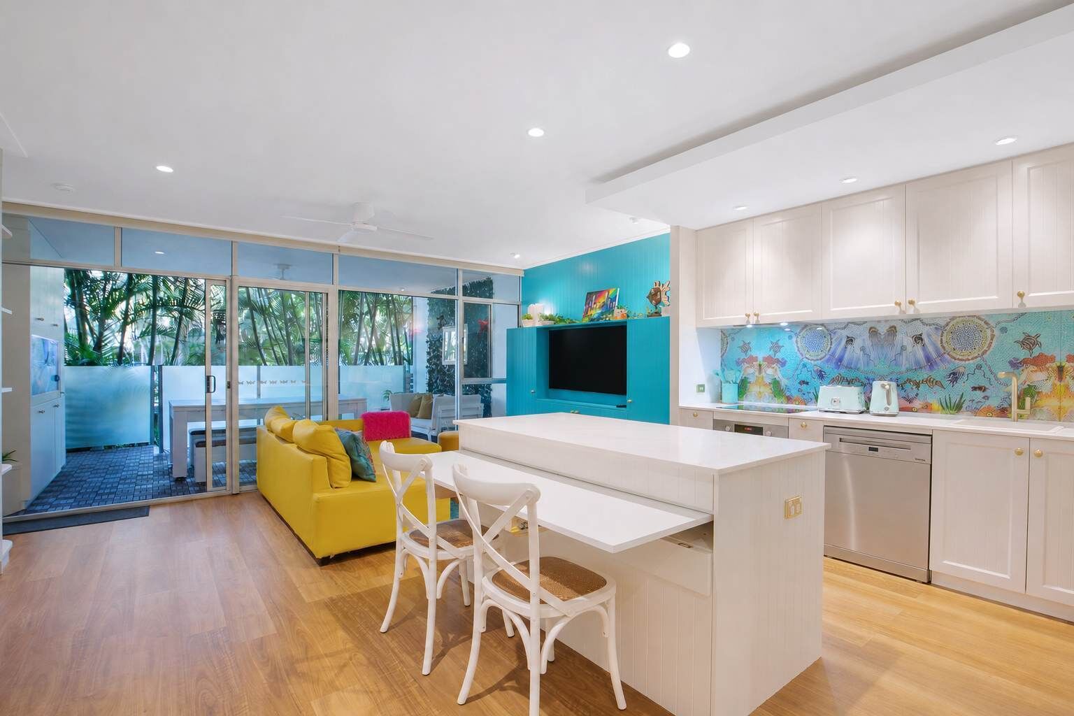 Une cuisine moderne avec un îlot central, un coin repas doté d’un canapé jaune et une grande fenêtre donnant sur un jardin tropical.