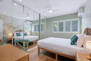 Ein Zimmer mit zwei Betten, einem Esstisch und einem Stuhl. Es gibt ein Fenster mit Jalousien, eine Klimaanlage und einen Deckenventilator.