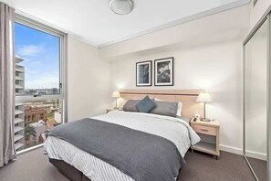Ein Hotelzimmer mit einem großen Bett, zwei Nachttischen mit Lampen, Blick auf die Stadt durch ein großes Fenster und zwei gerahmte Bilder an der Wand.