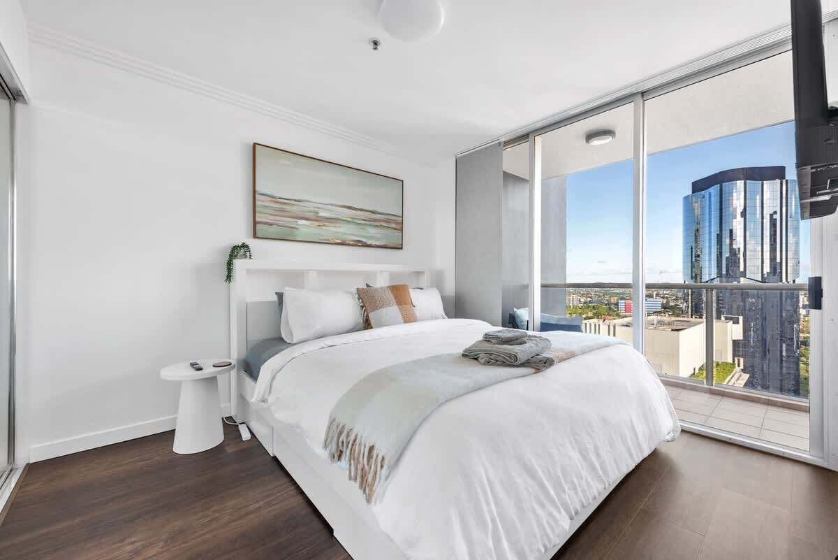 A modern bedroom with a large bed, a bedside table, a nightstand, and a city view through large windows.
