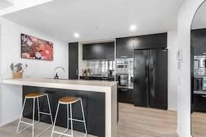 A modern kitchen with black cabinetry, a central island with stools, stainless steel appliances, and a decorative floral painting on the wall. - South Bank Sensational! ~ 2bed/2bath/1car/pool! (Brisbane)