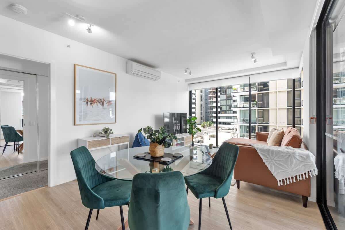 A modern living room with a glass dining table, teal chairs, a brown sofa, and a large window with a city view.