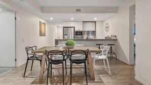 A modern kitchen with white cabinets, a central island with a plant, and a dining area with a wooden table and chairs. - South Bank Stunner! ~ 3 Bed/2 Bath/1car/views (Brisbane)