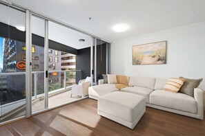 A modern living room with a sectional sofa, a glass door leading to a balcony, and a painting on the wall. - Urban Retreat 1bed-1bath-study-balcony-1car (Brisbane)