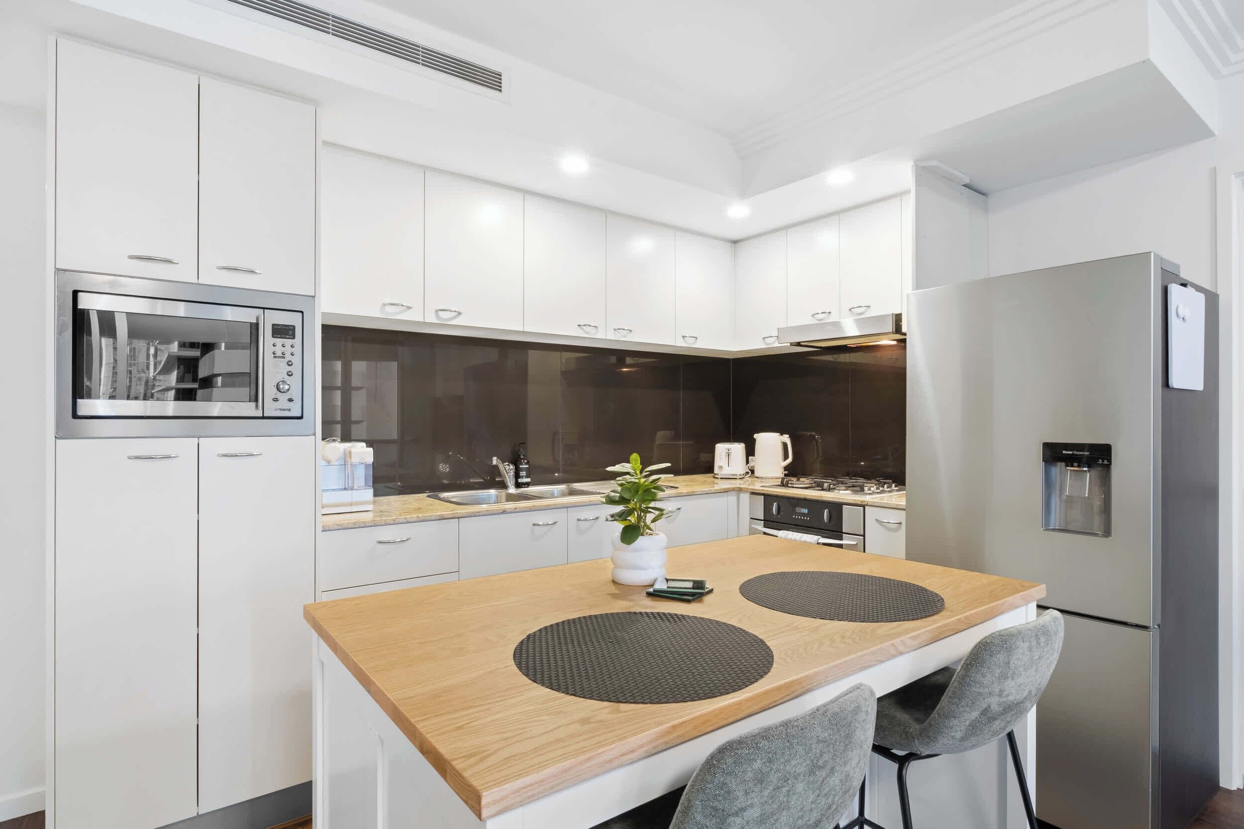 A modern kitchen with white cabinets, a stainless steel oven, a refrigerator, and a wooden island with two chairs.