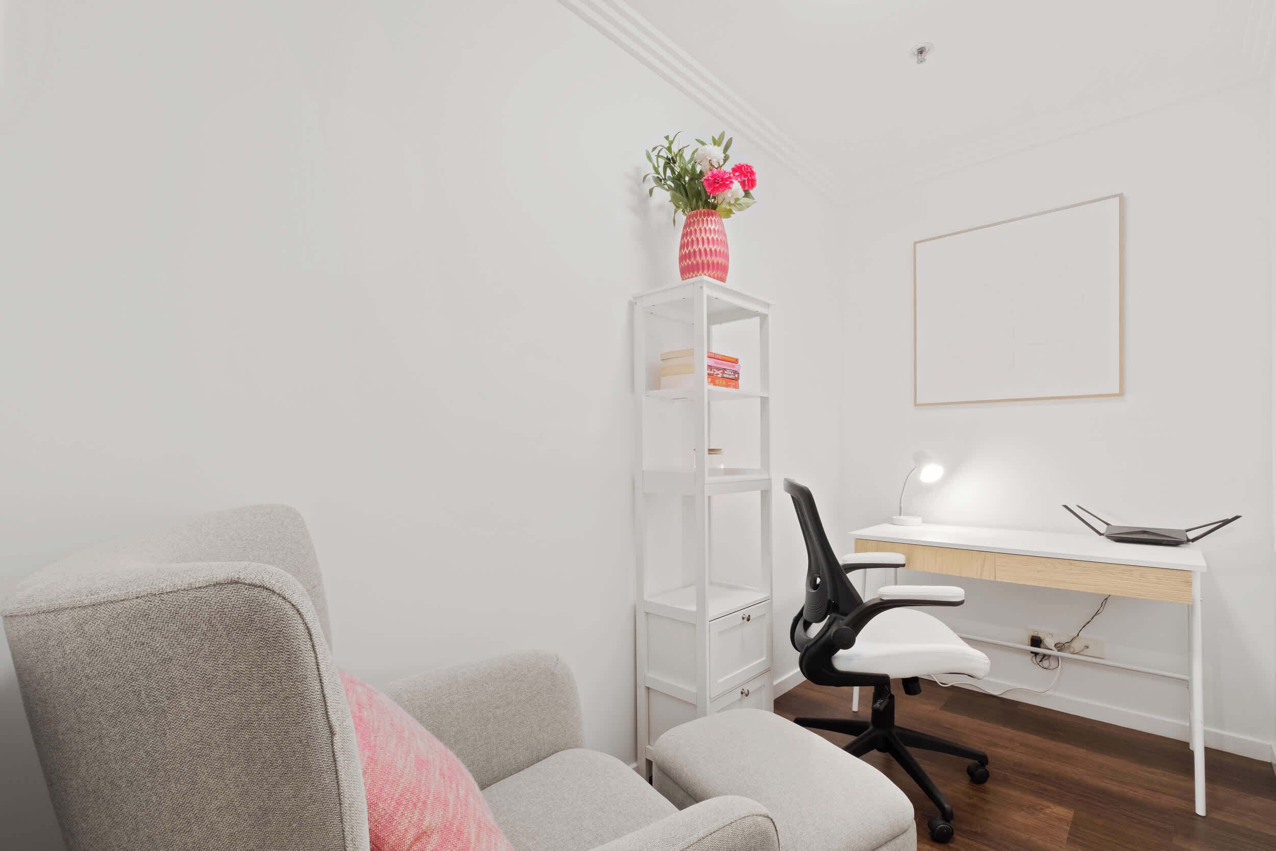 A room with a grey sofa, a white desk with a computer, a black office chair, and a white shelving unit with a vase of flowers.