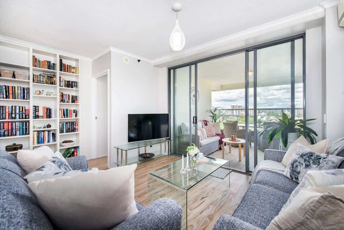 A modern living room with a sofa, a glass coffee table, a flat-screen TV, a bookshelf, and a balcony with a view.