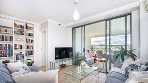 A modern living room with a sofa, a glass coffee table, a flat-screen TV, a bookshelf, and a balcony with a view. - Spectacular! 2bed, 1bath, 1car, Views ~ CBD (Brisbane)