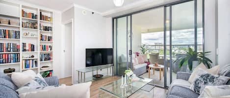 A modern living room with a sofa, a glass coffee table, a flat-screen TV, a bookshelf, and a balcony with a view.