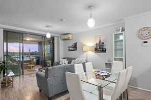A modern living room with a grey sofa, white chairs, a glass dining table, and a cityscape view through large glass doors. - Spectacular! 2bed, 1bath, 1car, Views ~ CBD (Brisbane)