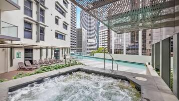 A rooftop pool with a hot tub, lounge chairs, and a modern building in the background.