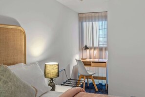 Habitació d'hotel amb llit, escriptori, cadira, làmpada i finestra amb cortines.