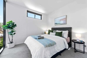 Ein modernes Schlafzimmer mit Bett, Nachttisch, Lampe, Fenster und einer Zimmerpflanze.