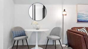 Ein Zimmer mit einem runden Spiegel, zwei Stühlen, einem weißen Tisch, einer Stehlampe, einem braunen Sofa und einem gerahmten Bild an der Wand.