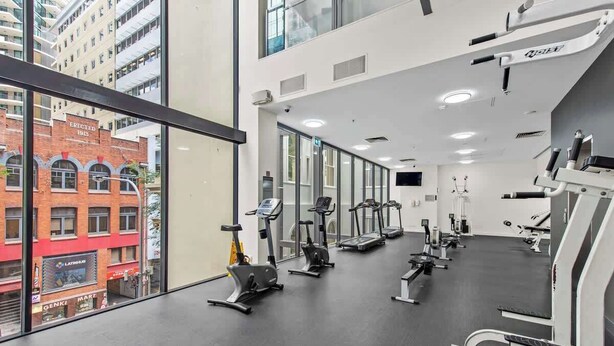 A gym with various exercise equipment, large windows, and a view of a red brick building. - City Centre! 1bed/1bath/study/1car/balcony ~ CBD (Brisbane)