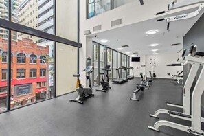 Une salle de sport équipée de divers appareils d’exercice, dotée de grandes fenêtres et offrant une vue sur un bâtiment en briques rouges.