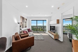 A modern living room with a sofa, TV, and large windows offering a city view. - Infinity Excellence!~1bed/1bath/office/1car~views! (Brisbane)